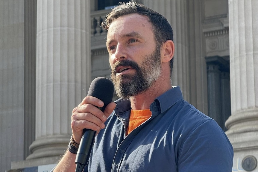 Warren Pickering holds a microphone and speaks on the steps of parliament