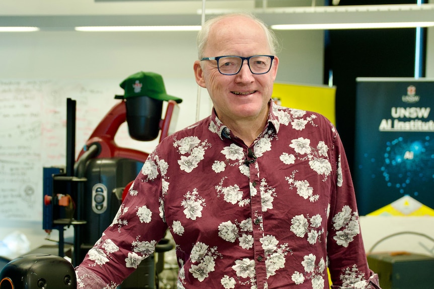 A grey haired man in black glasses smiles at the camera. He wears a patterned marron and white shirt.