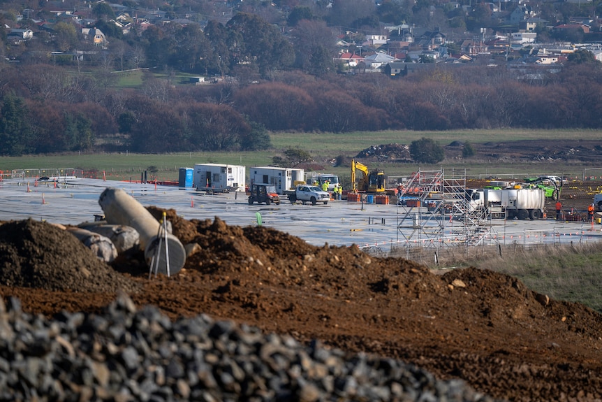 Piles of rocky dirt foregounds a construction site with yellow machinery in a valley dotted with homes.