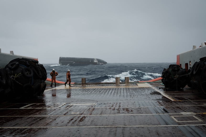 Two crew walk the deck of a ship in rough seas with a space rocket floating in water the background