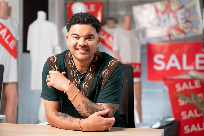 Guy Sebastian sits behind a table smiling. Behind him are shop mannequins and sale signs