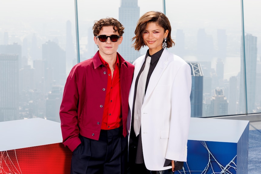 Tom Holland - a white man with curly hair, sunglasses, arm around Zendaya - a mixed Black woman white blazer, tie, short hair
