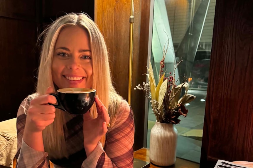A woman with long blonde hair holds up a coffee cup in a cafe.