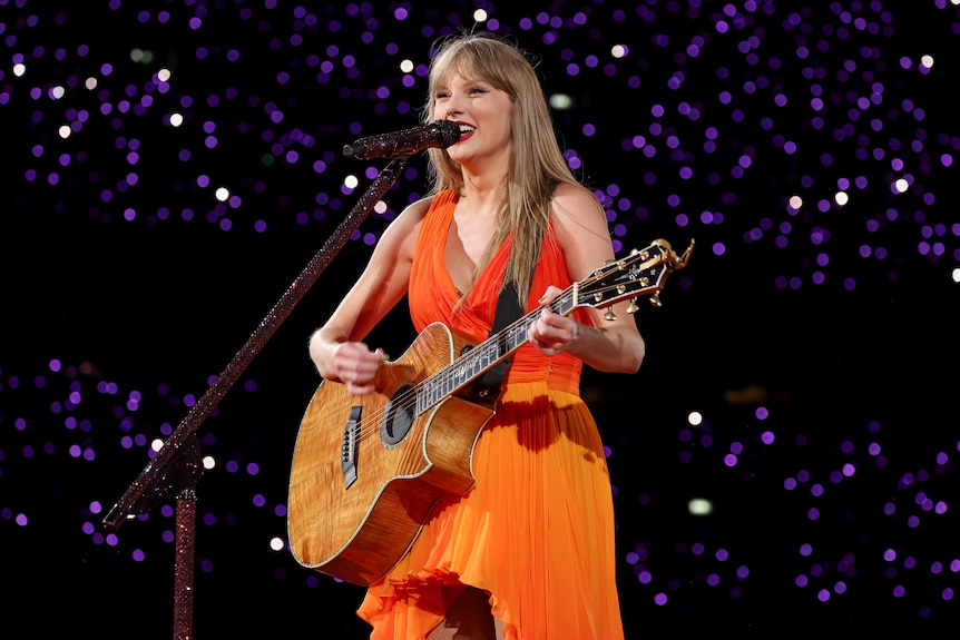 Taylor Swift smiles as she plays guitar on stage in an orange dress.