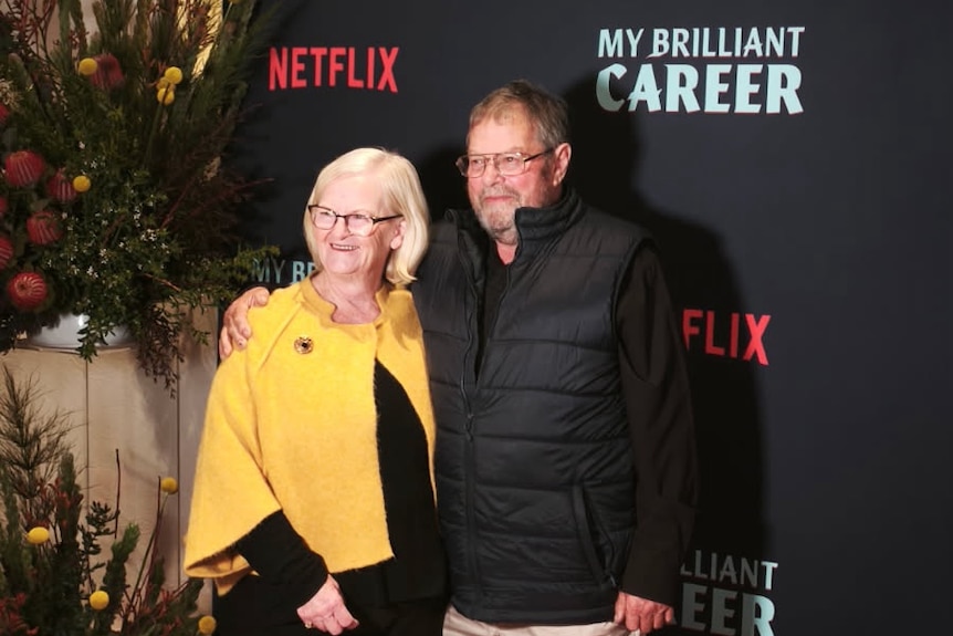 A couple smiles at a press event; they stand on a red carpet in front of a backdrop reading 'Netlix', 'My Brilliant Career'.