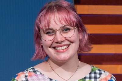 A headshot of a woman with pink hair and glasses smiling.