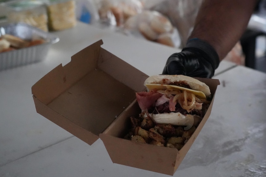 An outstretched hand holding out a takeaway box with a burger and potatoes