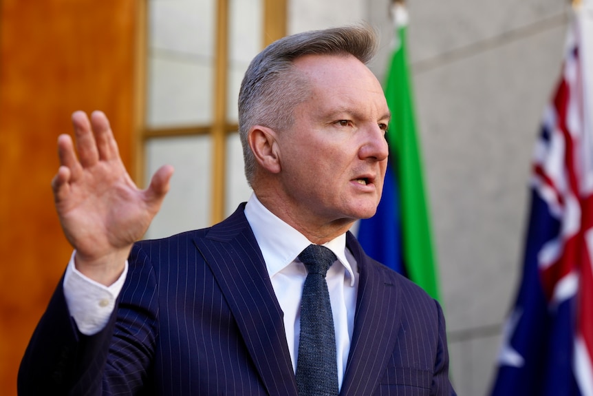 A middle-aged man with greying hair wears a suit and gestures as he speaks in front of a large building.