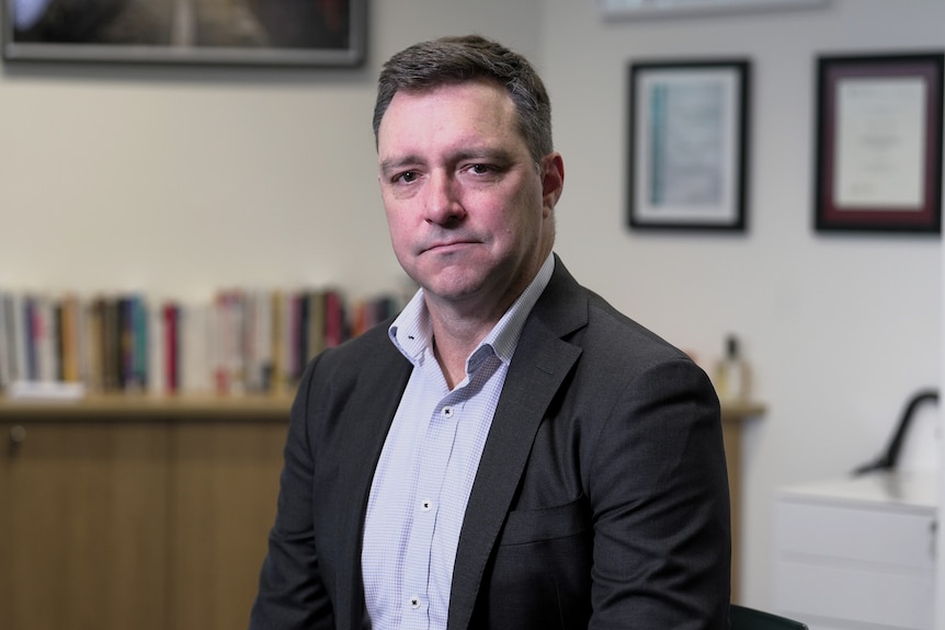 A middle aged white man with short brown hair sitting in an office in a dark suit