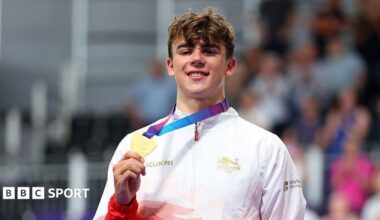 A smiling Henry Hobbs wearing a red and white Team England trackshuit shows his gold medal to the camera