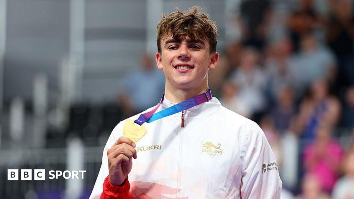 A smiling Henry Hobbs wearing a red and white Team England trackshuit shows his gold medal to the camera