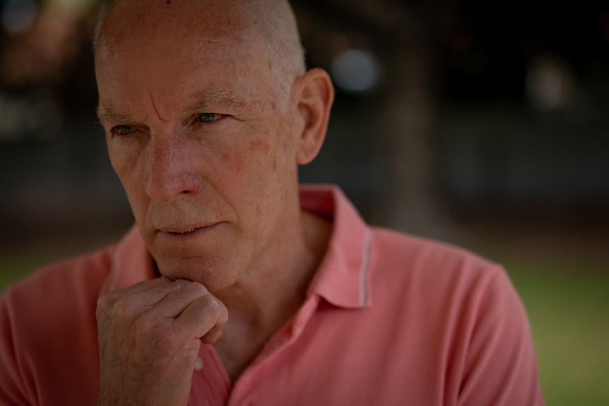 An older man wearing a salmon coloured shirt and a serious expression rests his chin in his hand.