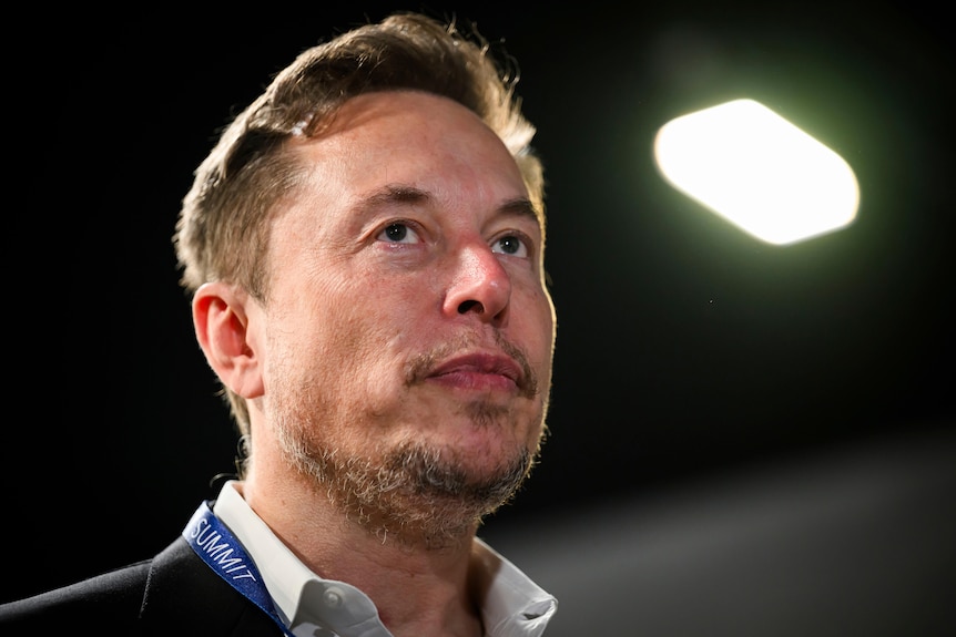 A man with short, dark hair and a light beard — Elon Musk — wears a lanyard and a blazer.