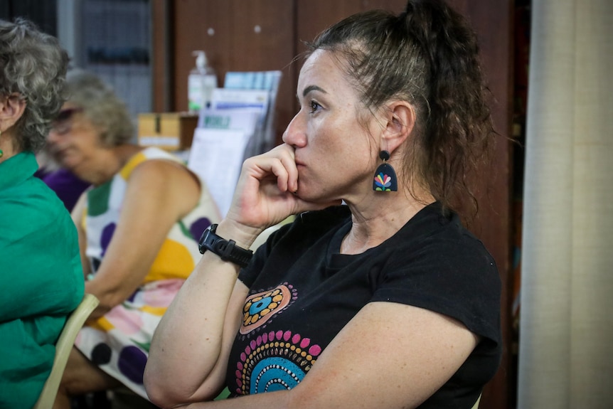 A mother with curly hair has a concerned facial expression, with her hand pressed to her jaw, as she sits in on a meeting.