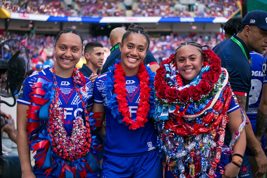 Three young Samoan girls wearing blue rugby league uniforms with lei around their necks smile at the camera