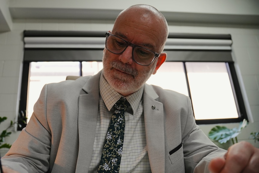 A bald man with salt and pepper beard and glasses poses for camera in his office. He is wearing glasses.