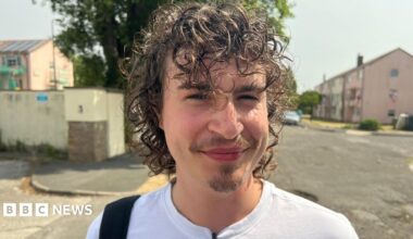 Archie Byron is smiling at the camera. the image is a head and shoulders shot of Archie. He has dark curly hair and a small beard on his chin. He is wearing a white T-shirt and has a bag over one shoulder. He is stood in an urban area with a block of flats in the background.