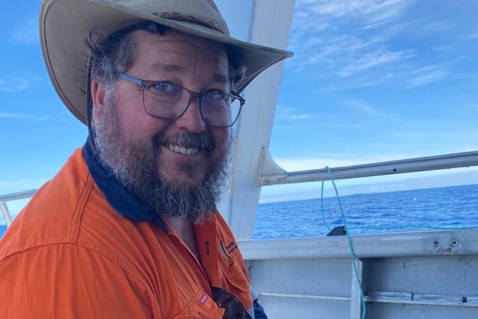 A man sitting on a boat, wearing an orange work shirt, smiling.