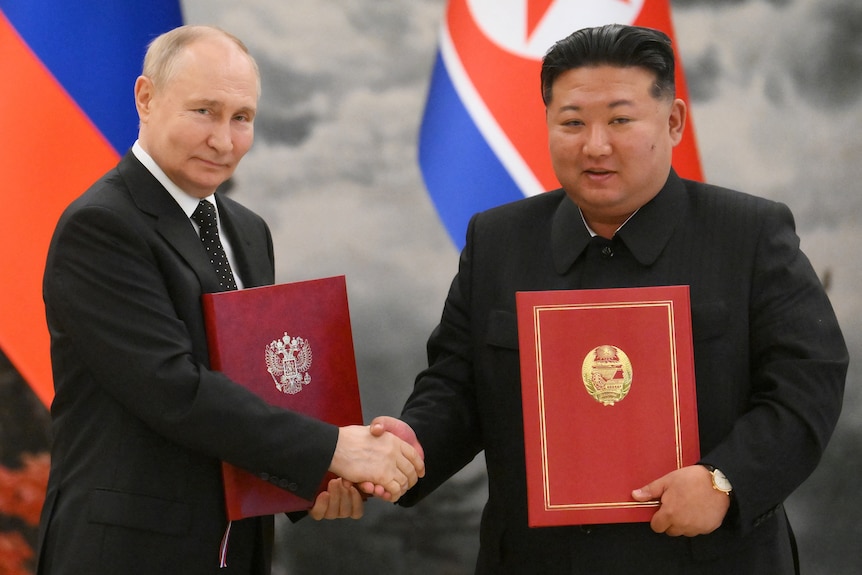 Two men are shaking hands and holding folders while standing in front of country flags.