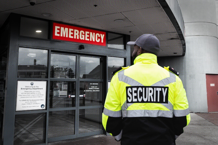 A security guard stands with his back to the camera in front of an emergency department entrance