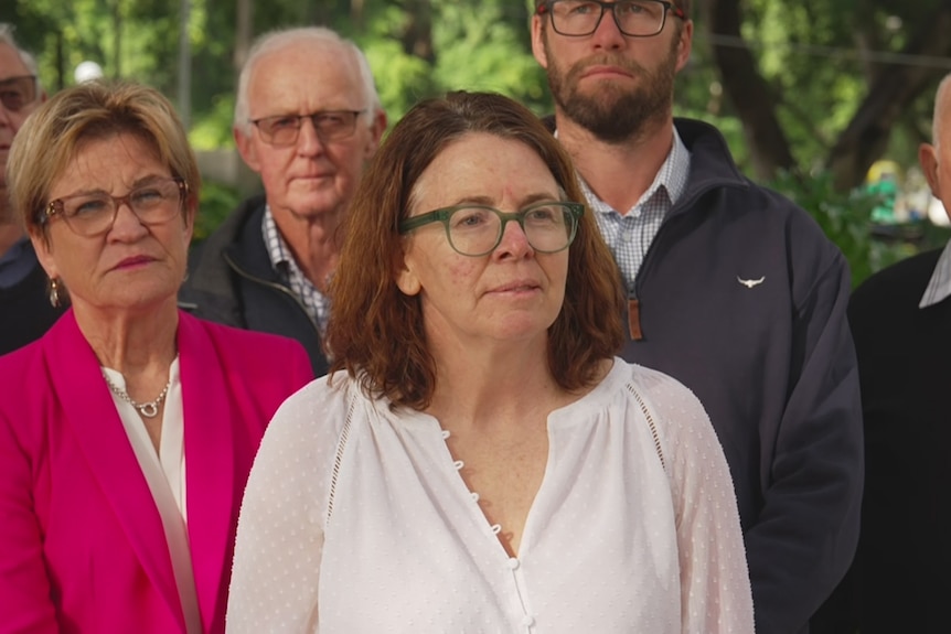 Sophie Baldwin, a woman with long brown hair who is wearing a white top, stands in front of a group of people.