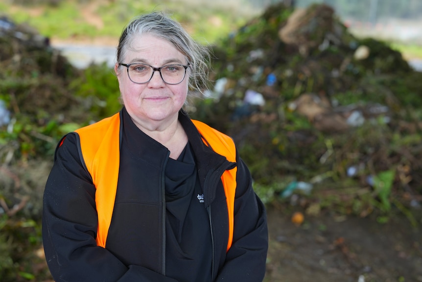 A woman with grey hair and glasses wearing a high vis vest stands in front of a pile of contaminated green waste