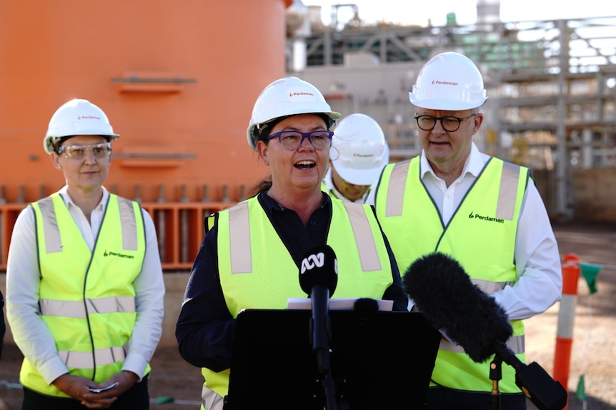 A dark-haired, middle-aged woman in glasses, high-vis and a hard hat speaks at an industrial site.