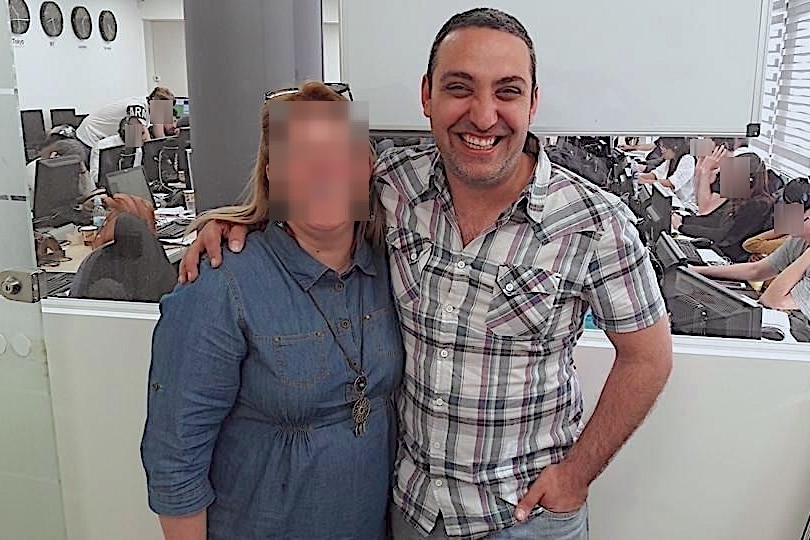 A middle aged man stands in a call centre, smiling beside a woman with her face blurred out