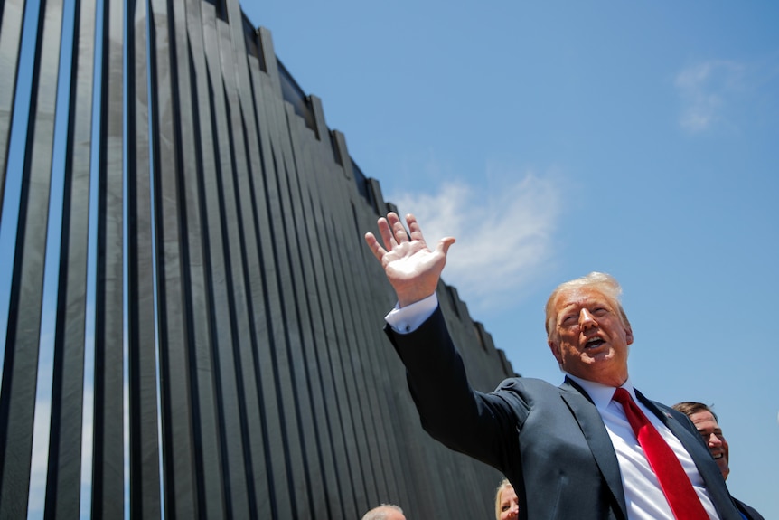 Donald Trump wears a black suit and red tie and stands by the thick bars of a border wall, about to sign a plaque.