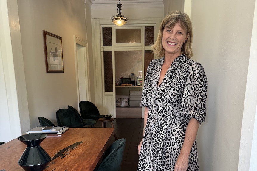 Woman in leopard print grey and black dress with shoulder length blonde hair smiles from inside restaurant.