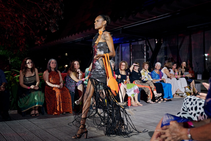 A tall woman wearing a black short dress with floor length tassels coming from the hem holding orange bag