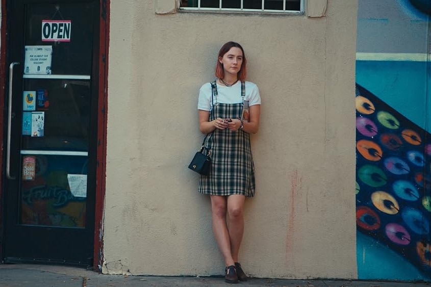 A scene from a film shows a young woman standing against a wall