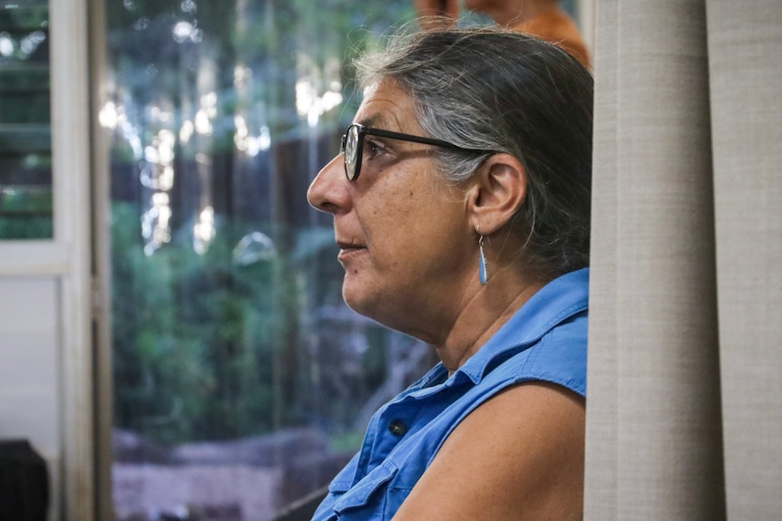 An older woman, grey hair, is seated inside a community hall, she faces the front of the room with a serious look on her face.