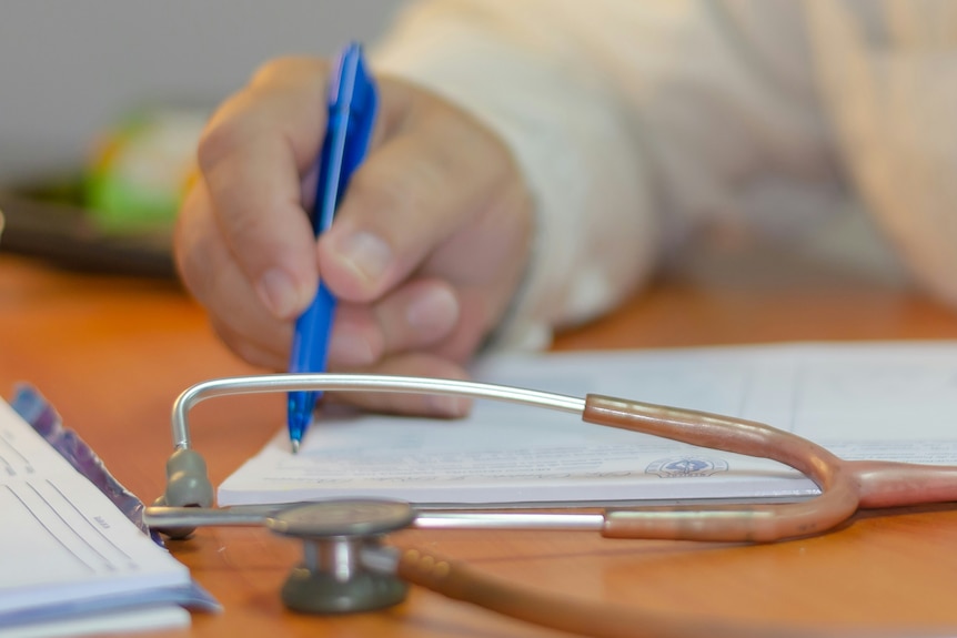 A female doctor writing in a note pad, with a stethoscope on the table.