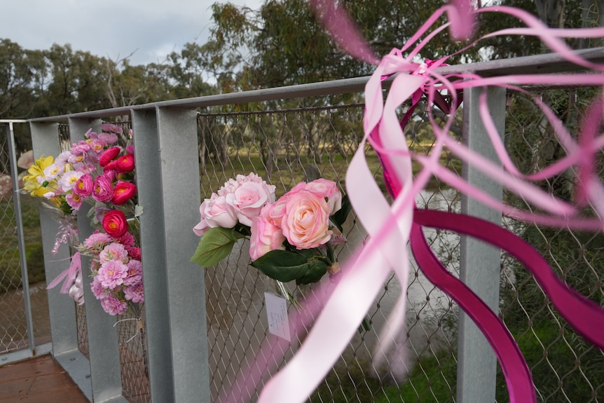 Half a dozen flower bouquettes zip tied to wire fencing at memorial in front of river, pink ribbons flowing across frame