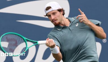 Toby Samuel, wearing a grey shirt and white backwards baseball cap, hits the ball with a green racquet