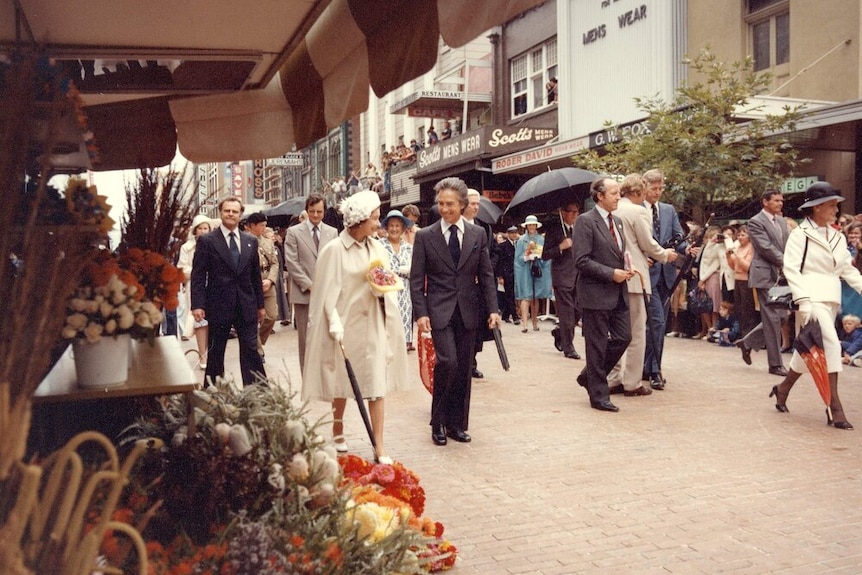 An archived image of the Queen wearing a white dress, walking in a shopping mall with a man in suit surrounded by crowd.