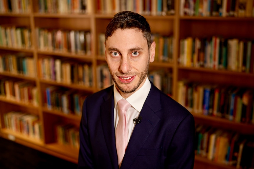 A man wearing a dark suit, white shirt and pink tie stands in a room in front of a bookshelf filled with books.