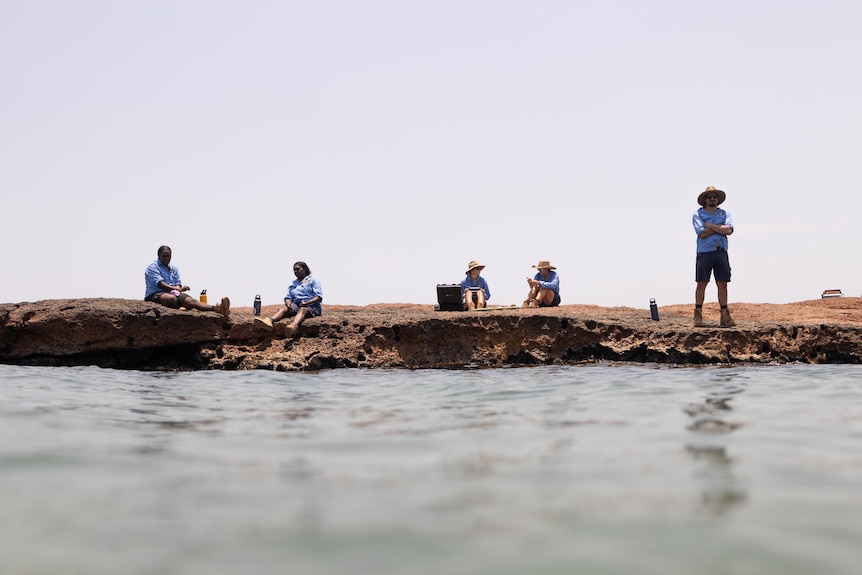 A group of researchers and Indigenous rangers standing on the shore of a beach, one looks out over the ocean.