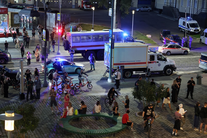 An night-time aerial image shows a busy street with multiple emergency vehicles and pedestrians and cyclists looking on