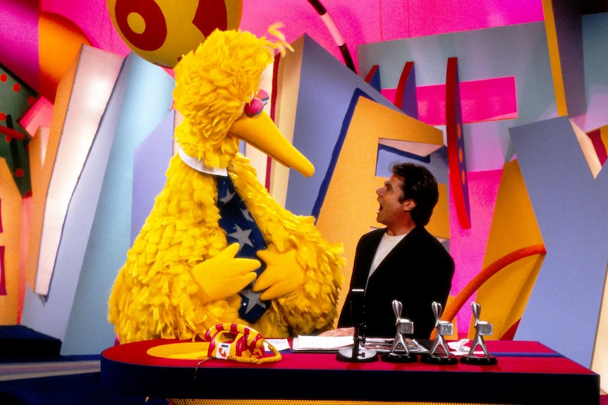 Big Bird - a costumed person wearing yellow feathers with long beak next to Daryl Somers a man with black hair, mouth open wide.