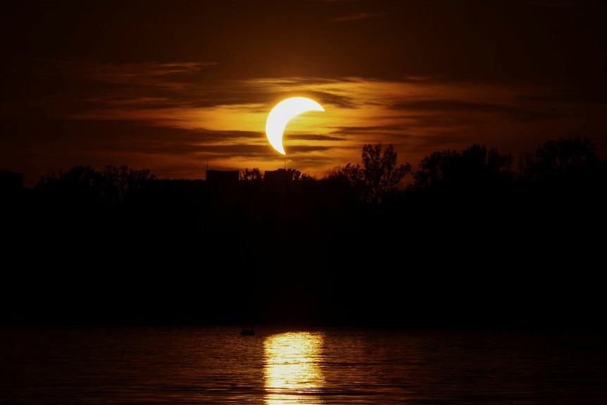 A partial solar eclipse shines over a lake.