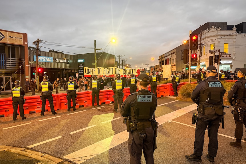 Police line an orange barrier that separates them from a large group of people with banners on dusk.