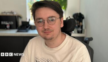 Jack is wearing glasses and a cream T-shirt. He is sitting in his wheelchair in the kitchen