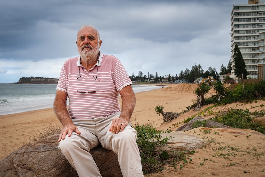A man in a striped red and white shirt sits on a rock on a beach - looking at the camera.