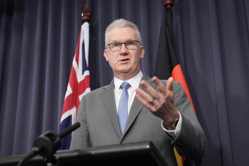 A clean-shaven man with grey hair and glasses, speaking at a lectern.