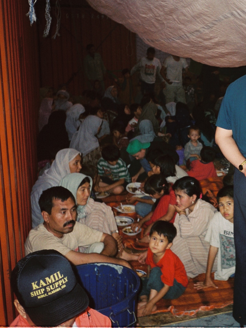 A crowd of people, including young children, stand and sit on the floor beneath a tarp.