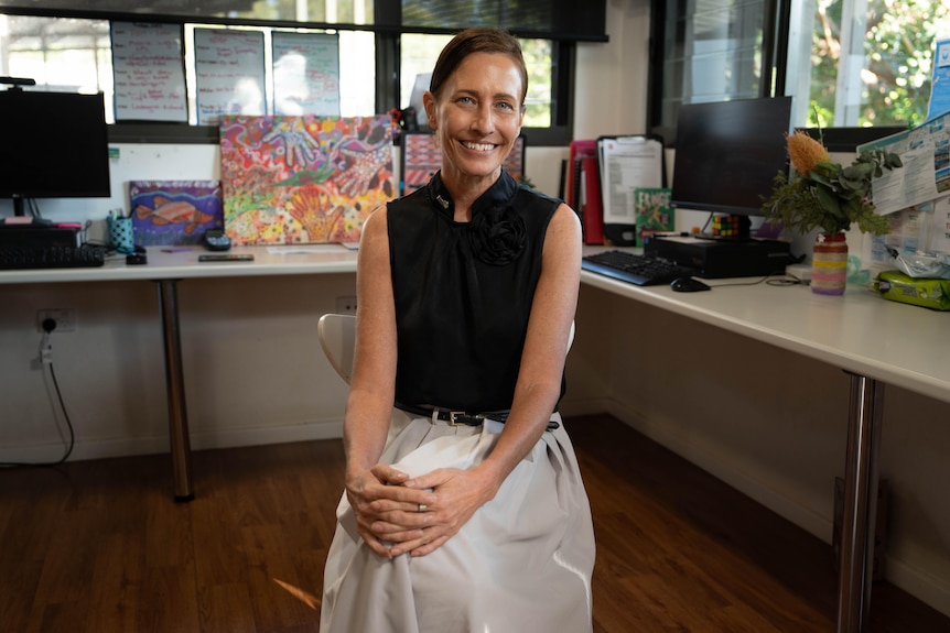 Rina sits inside an office, the woman has light eyes, brunette hair, and is looking at the camera smiling.