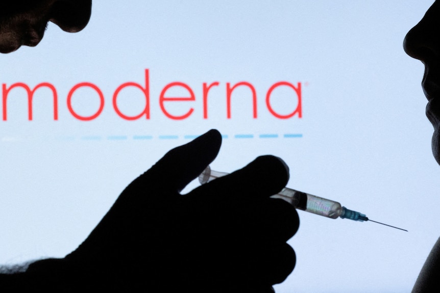 A person holding a syringe up to another person with the Moderna logo illuminated behind them.