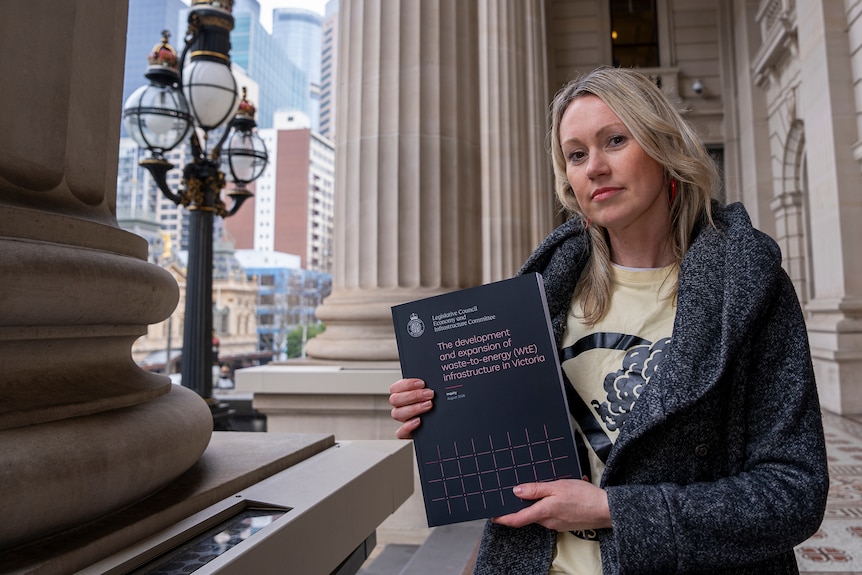 A woman with blonde hair in a black coat holds up a black book while standing at Parliament House.
